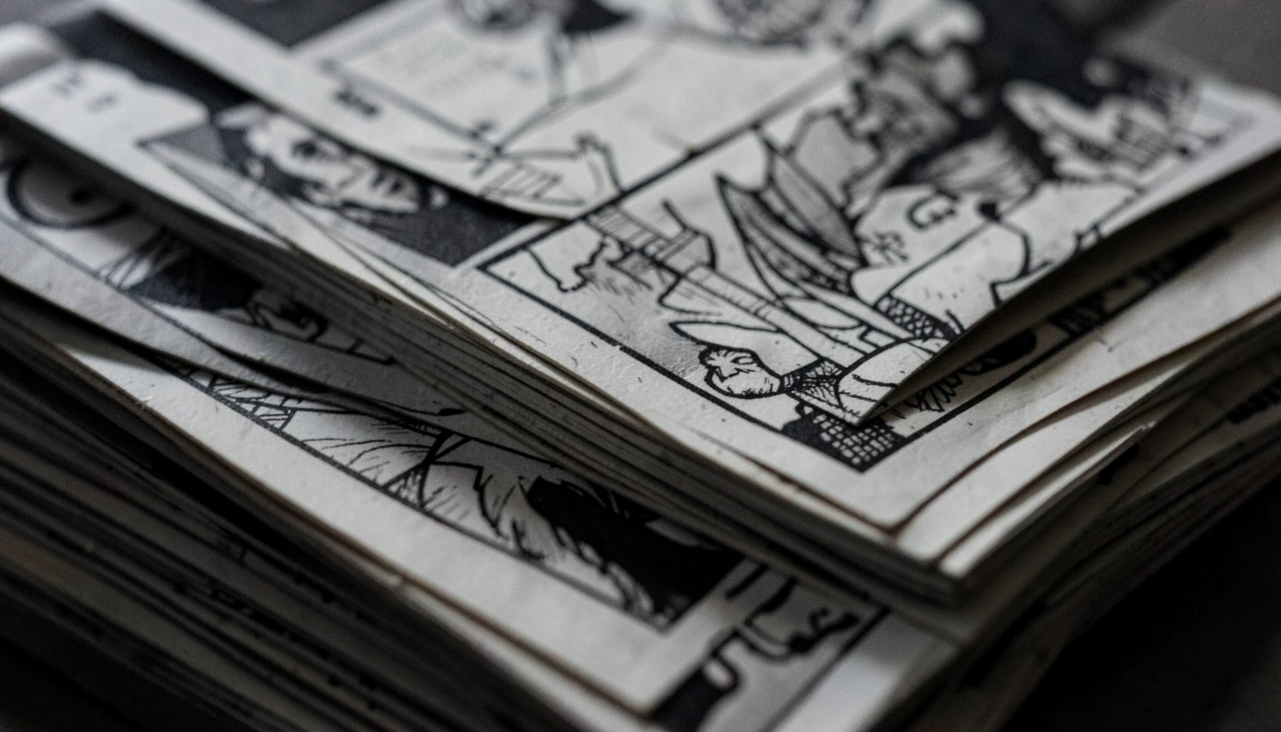 A moody, desaturated close-up of stacked comic book pages with black-and-white line art visible, dark grays and muted off-whites dominating the scene