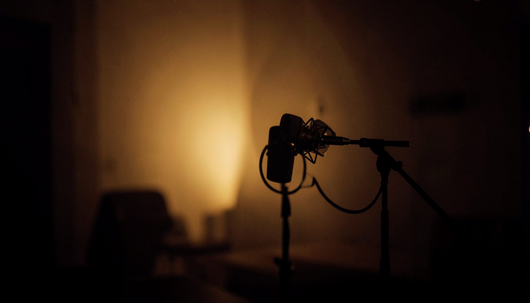 A moody, dimly lit interior with warm amber tones, a single microphone on a stand silhouetted against soft backlight