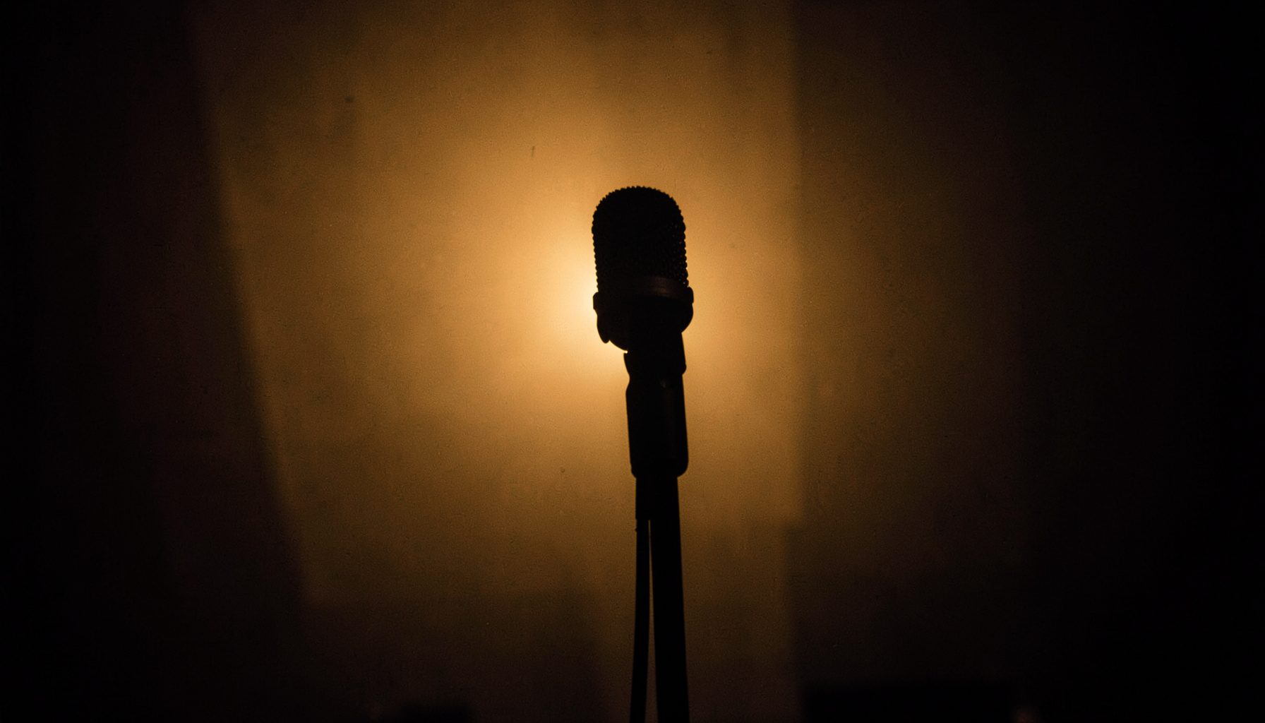 A moody, dimly lit interior with warm amber tones, a single microphone on a stand silhouetted against soft backlight