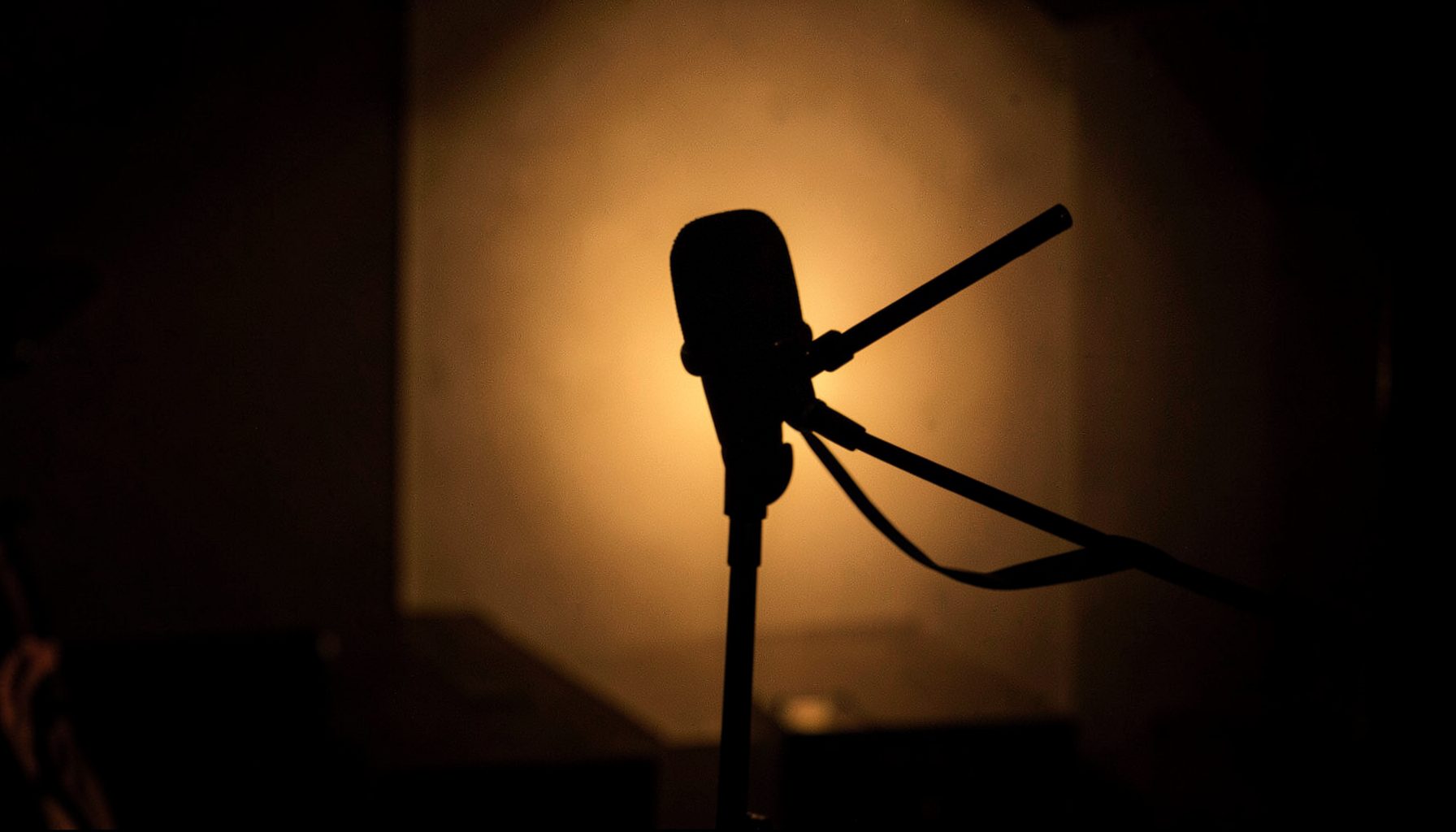 A moody, dimly lit interior with warm amber tones, a single microphone on a stand silhouetted against soft backlight, suggesting an interview or behind-the-scenes moment
